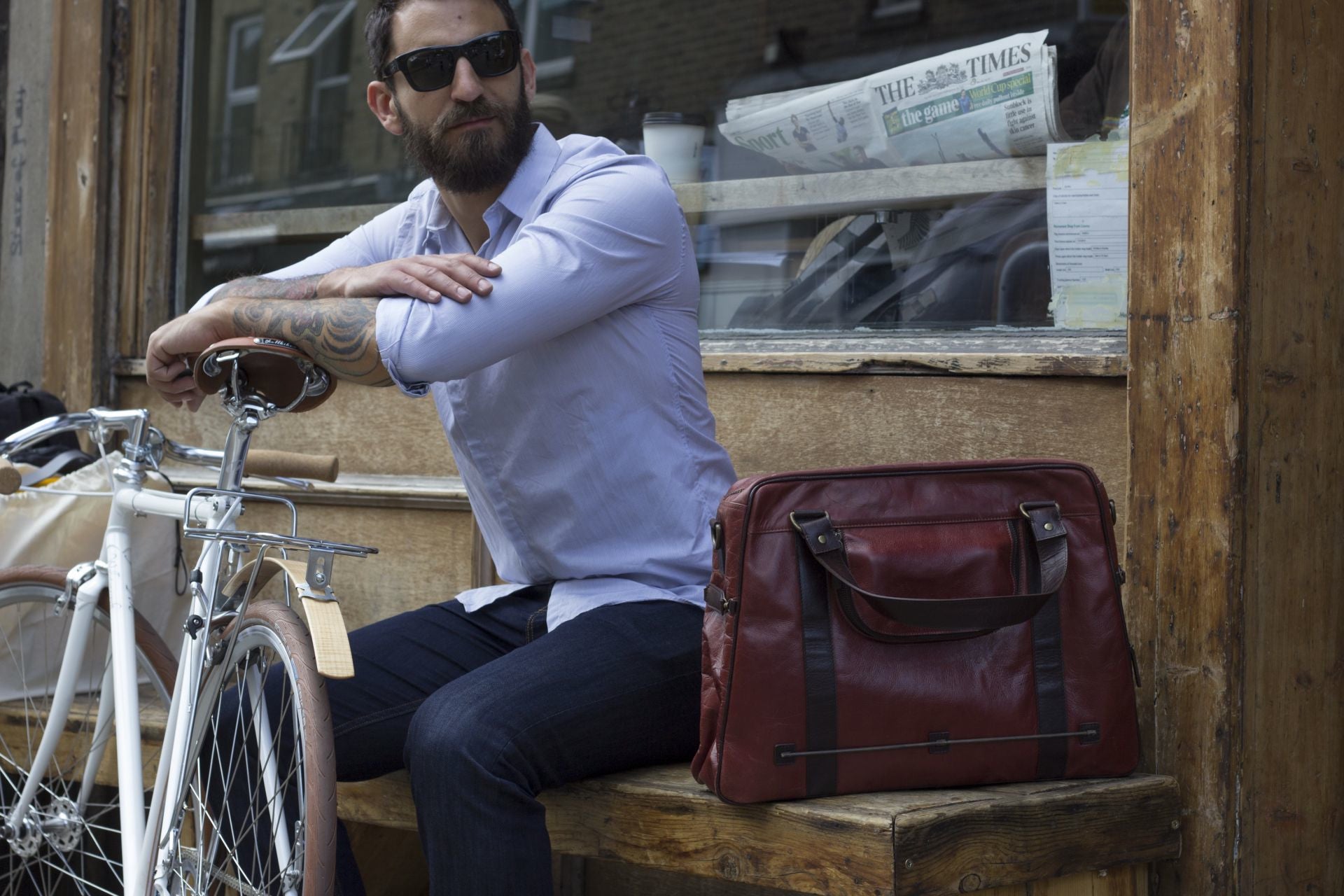 Hill and Ellis Duke Cherry Red Leather Satchel with model sitting on a bench beside a bicycle